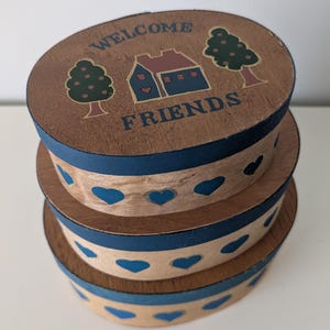 May include: A stack of three wooden boxes with a rustic design. The top box lid features the words "WELCOME FRIENDS" with a house and tree illustration. The sides of the boxes are decorated with blue hearts.