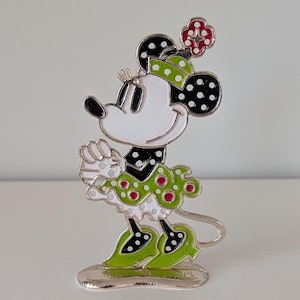 May include: A Minnie Mouse figurine in a standing pose. The figure has a white face, black ears with white polka dots, and a green and white polka dot dress. The figure is accented with red polka dots and a red flower on her head. The base is silver.