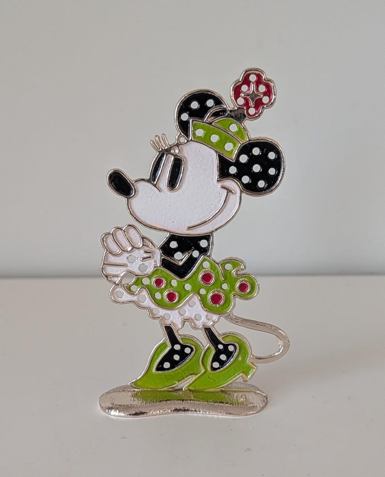 May include: A Minnie Mouse figurine in a standing pose. The figure has a white face, black ears with white polka dots, and a green and white polka dot dress. The figure is accented with red polka dots and a red flower on her head. The base is silver.