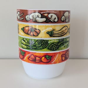 May include: A white ceramic bowl with colorful bands featuring illustrations of vegetables. The bands display mushrooms, squash, asparagus, and tomatoes, with the corresponding names written above each image. The bowl is stacked with the vegetables in descending order.