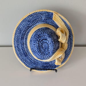 May include: A ceramic decorative plate shaped like a wide-brimmed straw hat. The hat features a textured blue woven pattern with a cream-colored brim edge and a matching cream-colored ribbon bow detail wrapped around the crown of the hat.