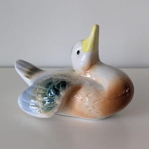 May include: A ceramic duck figurine with a white head and yellow beak, brown body, and blue and green wings. The duck is in a resting position with its head raised. The figurine is a decorative item.
