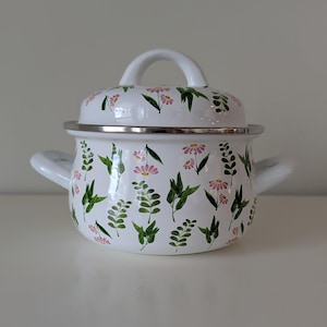 May include: A white enamel pot with a matching lid, featuring a repeating pattern of small pink flowers and green leaves. The pot has two side handles and a rounded lid with a central handle, all finished with a smooth, glossy white surface.