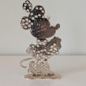 May include: A silver-colored Minnie Mouse figurine with a polka-dot design. The figurine has a small base and the text "WALT DISNEY PRODUCTIONS" is visible on the back. The figure is a collectible item.