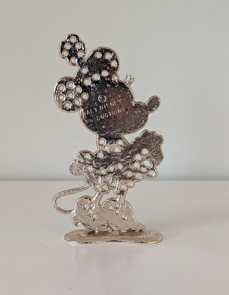 May include: A silver-colored Minnie Mouse figurine with a polka-dot design. The figurine has a small base and the text "WALT DISNEY PRODUCTIONS" is visible on the back. The figure is a collectible item.