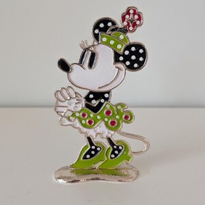 May include: A Minnie Mouse figurine with a white face, black ears with white polka dots, and a green and red polka dot outfit. The figurine has a silver base and is standing on a white surface.
