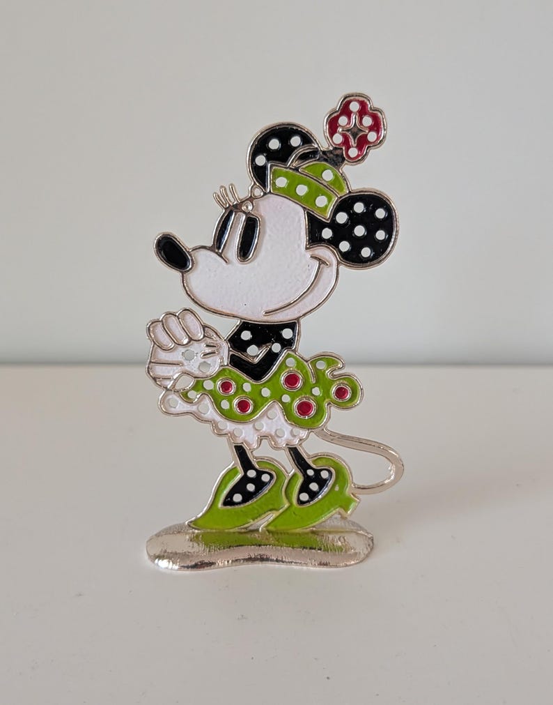 May include: A Minnie Mouse figurine with a white face, black ears with white polka dots, and a green and red polka dot outfit. The figurine has a silver base and is standing on a white surface.