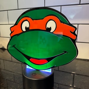 May include: A green Teenage Mutant Ninja Turtles character face night light with an orange mask and a red mouth. The light is on a black base with a power button.