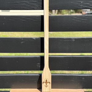 May include: A light-colored wooden paddle with a long handle and a flat, paddle-shaped end. The end features a laser-etched design of a crawfish. The paddle is displayed against a black slatted background and a wooden shelf.