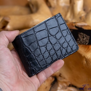 May include: A black bifold wallet featuring a textured crocodile skin pattern. The wallet has visible stitching along the edges and a rectangular shape. A black credit card with gold detailing is partially visible tucked into the wallet slot.