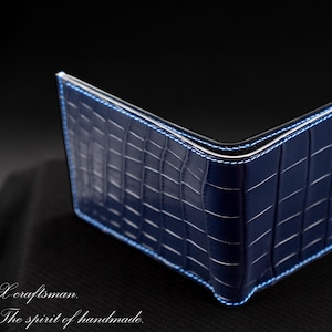 Könnte beinhalten: Ein dunkelblaues Bifold-Portemonnaie mit glänzender Krokodilleder-Textur und hellblauen Nähten an den Kanten. Das Portemonnaie ist V-förmig geöffnet. Der Text "X craftsman" und "The spirit of handmade" ist in weißer Schrift unten links gedruckt.