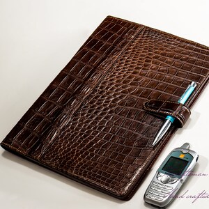 May include: A brown leather folder with a crocodile pattern. The folder has a pen holder and a slot for a phone. The text "hand crafted with passion" is visible in the bottom right corner.