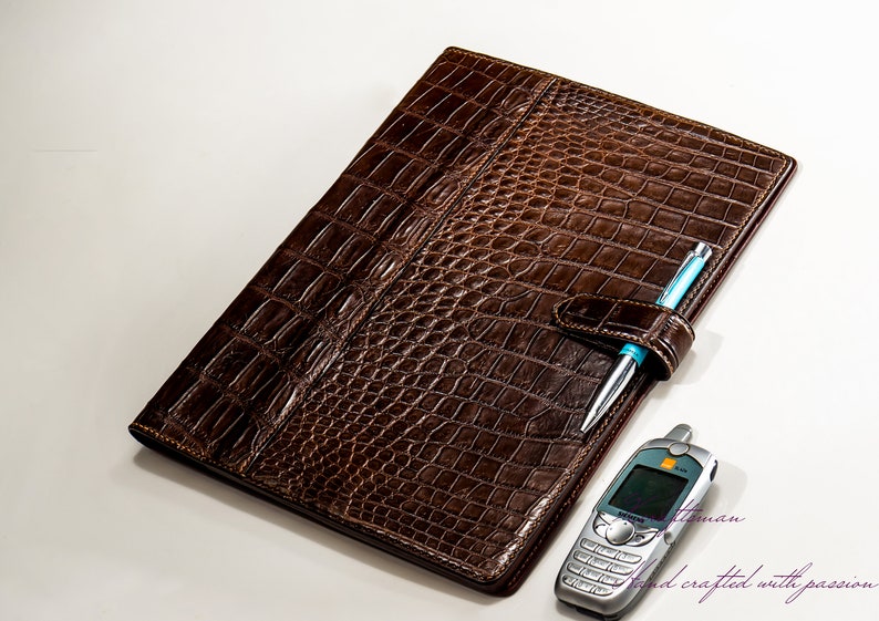 May include: A brown leather folder with a crocodile pattern. The folder has a pen holder and a slot for a phone. The text "hand crafted with passion" is visible in the bottom right corner.