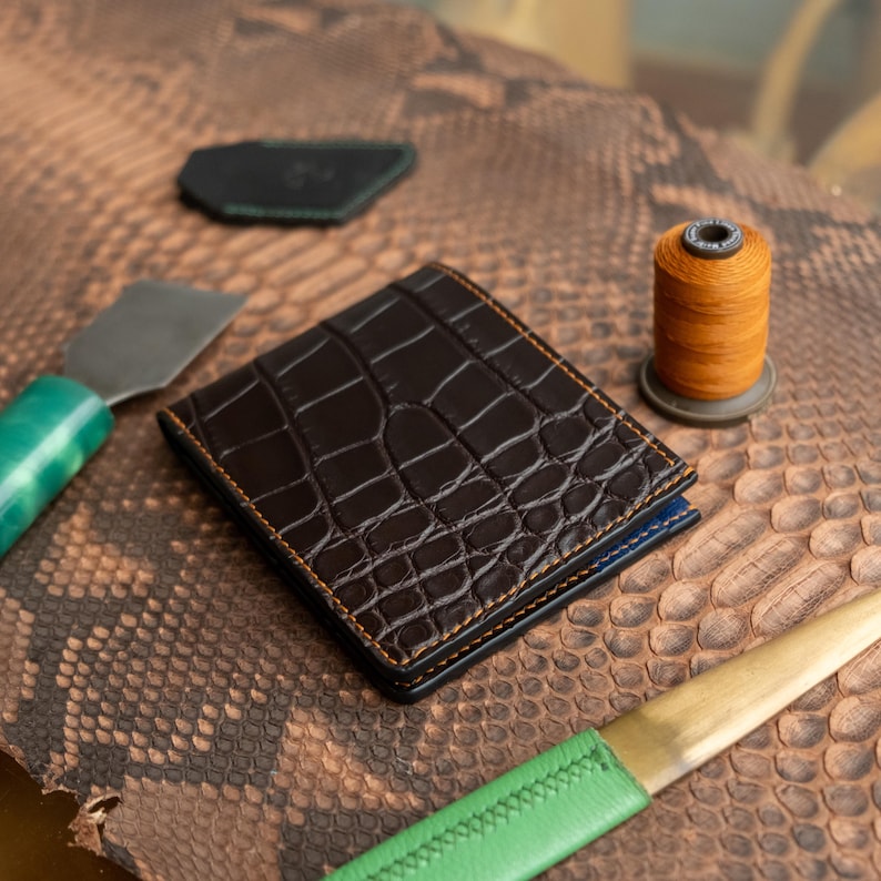 May include: A brown, crocodile-textured leather wallet with orange stitching. A spool of orange thread, a green-handled tool, and a black leather piece are also visible. The wallet has a blue interior.