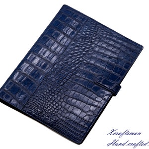 May include: A dark blue leather folder with a crocodile pattern. The folder has a flap closure with a strap and buckle. The text "Keraftsman Hand crafted with passion." is printed on the bottom right corner of the image.