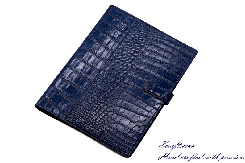 May include: A dark blue leather folder with a crocodile pattern. The folder has a flap closure with a strap and buckle. The text "Keraftsman Hand crafted with passion." is printed on the bottom right corner of the image.