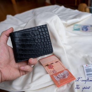 Puede incluir: Una cartera de cuero negra con un patr&oacute;n de cocodrilo. La cartera est&aacute; siendo sostenida en una mano.