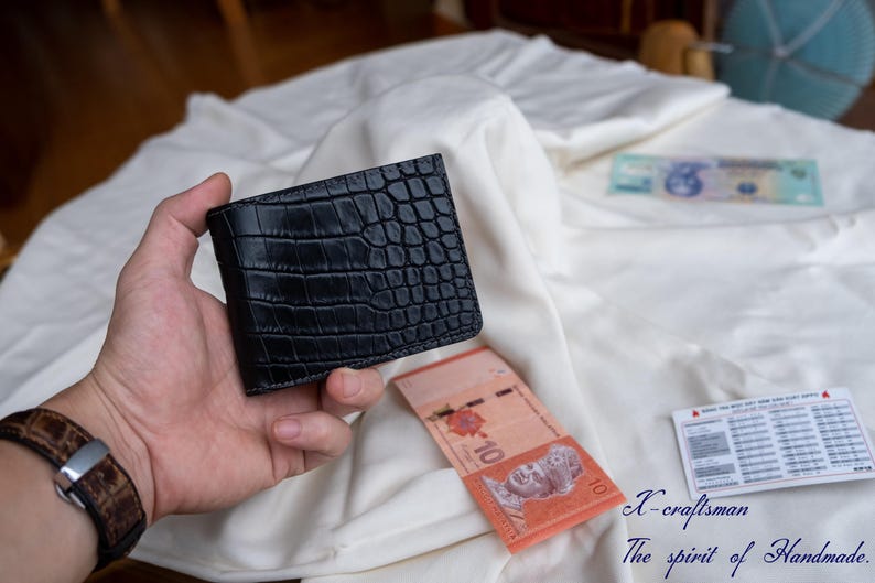 Puede incluir: Una cartera de cuero negra con un patr&oacute;n de cocodrilo. La cartera est&aacute; siendo sostenida en una mano.