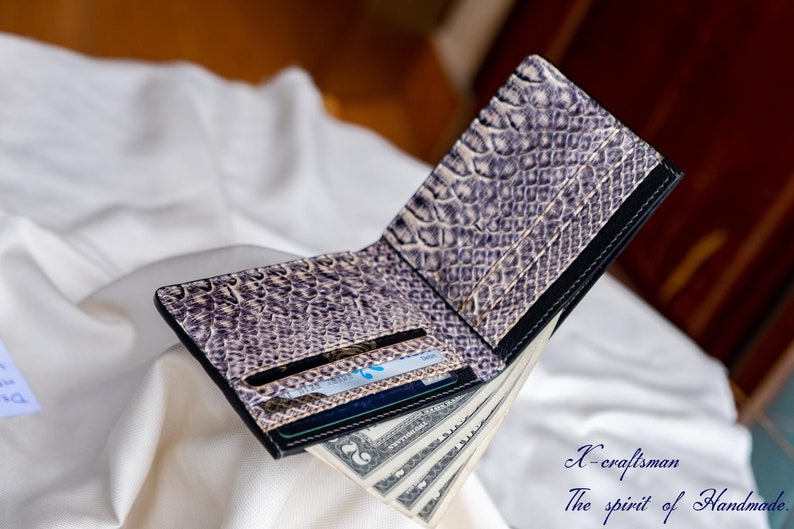 Puede incluir: Una cartera de cuero negro con un patr&oacute;n de piel de serpiente y una tarjeta de cr&eacute;dito y dinero en efectivo en su interior. La cartera est&aacute; abierta y el interior es visible. La cartera est&aacute; sobre una superficie blanca.