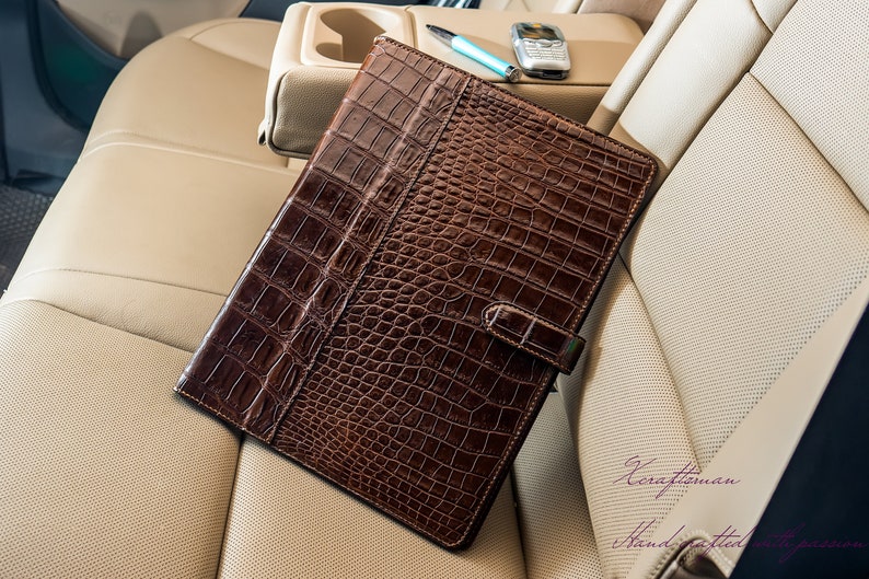 May include: A brown leather folder with a crocodile pattern. The folder is closed and has a strap with a buckle. The folder is sitting on a beige leather seat.