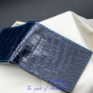 May include: A dark blue leather wallet with a crocodile pattern and blue stitching. The wallet is folded in half and lying on a white surface.