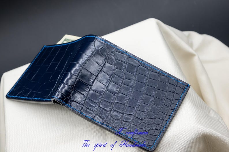 May include: A dark blue leather wallet with a crocodile pattern and blue stitching. The wallet is folded in half and lying on a white surface.