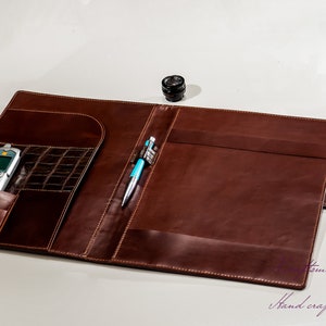 May include: A brown leather portfolio with a pen and a cell phone inside. The portfolio has a flap that closes with a strap and a brass button. The text "Hand crafted with passion" is visible at the bottom of the image.