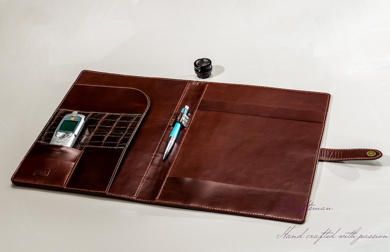 May include: A brown leather portfolio with a pen and a cell phone inside. The portfolio has a flap that closes with a strap and a brass button. The text "Hand crafted with passion" is visible at the bottom of the image.