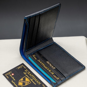May include: A black leather wallet with blue stitching and a blue interior. The wallet is open and has several card slots. The wallet is sitting on a white surface. The text "XCRAFTSMAN The spirit of Handmade." is visible on a business card.