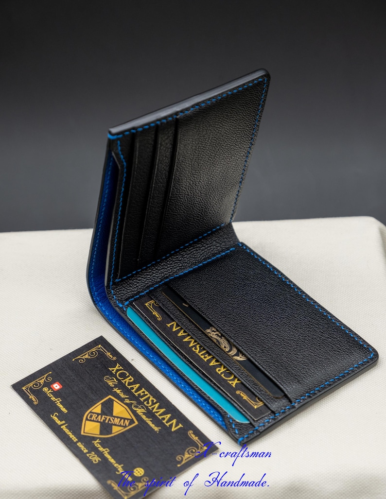 May include: A black leather wallet with blue stitching and a blue interior. The wallet is open and has several card slots. The wallet is sitting on a white surface. The text "XCRAFTSMAN The spirit of Handmade." is visible on a business card.