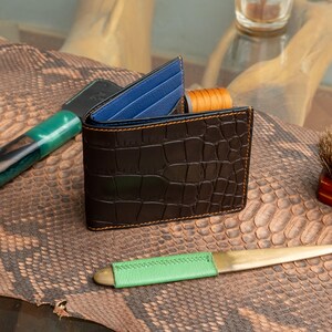 May include: A black leather wallet with a crocodile texture, featuring blue card slots and orange stitching. The wallet is open, displaying the interior. The background includes leather tools and a snakeskin-like surface.