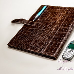May include: A brown leather folder with a crocodile pattern and a blue pen sticking out of the top. A silver cell phone is lying on the white surface next to the folder. The text "Craftsman" and "Hand crafted with passion" is written in purple on the white surface.