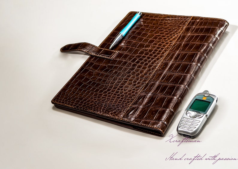May include: A brown leather folder with a crocodile pattern and a blue pen sticking out of the top. A silver cell phone is lying on the white surface next to the folder. The text "Craftsman" and "Hand crafted with passion" is written in purple on the white surface.