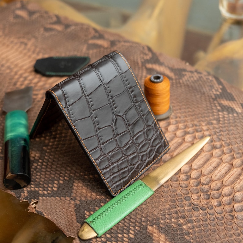 May include: A brown leather wallet with a crocodile pattern, featuring orange stitching. A green-handled letter opener and a spool of orange thread are also visible, suggesting a leathercraft theme.