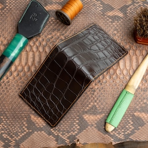 May include: A dark brown leather wallet with a crocodile skin texture, featuring orange stitching. The wallet is surrounded by leather tools and a spool of thread, suggesting a handcrafted leather goods theme.
