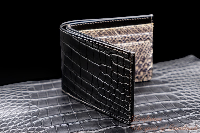 Puede incluir: Una cartera de piel de cocodrilo negra con interior de piel de serpiente. La cartera est&aacute; abierta y sobre una superficie de piel de cocodrilo.