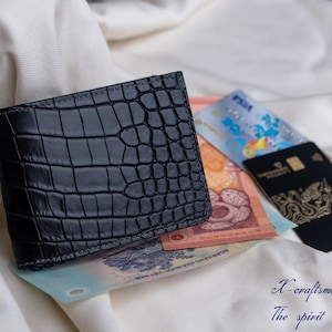 Puede incluir: Una cartera de piel de cocodrilo negra con una superficie texturizada. La cartera est&aacute; abierta y tiene varios billetes y tarjetas en su interior.