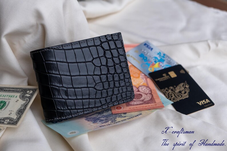 Puede incluir: Una cartera de piel de cocodrilo negra con una superficie texturizada. La cartera est&aacute; abierta y tiene varios billetes y tarjetas en su interior.
