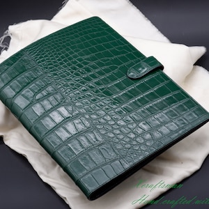 May include: A dark green leather binder with a crocodile pattern. The binder has a strap closure and is lying on a white fabric surface.