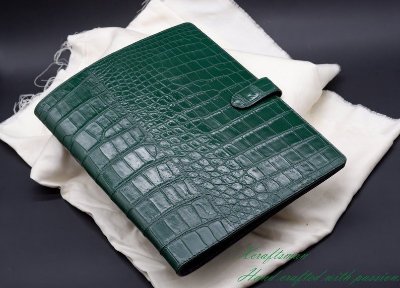 May include: A dark green leather binder with a crocodile pattern. The binder has a strap closure and is lying on a white fabric surface.