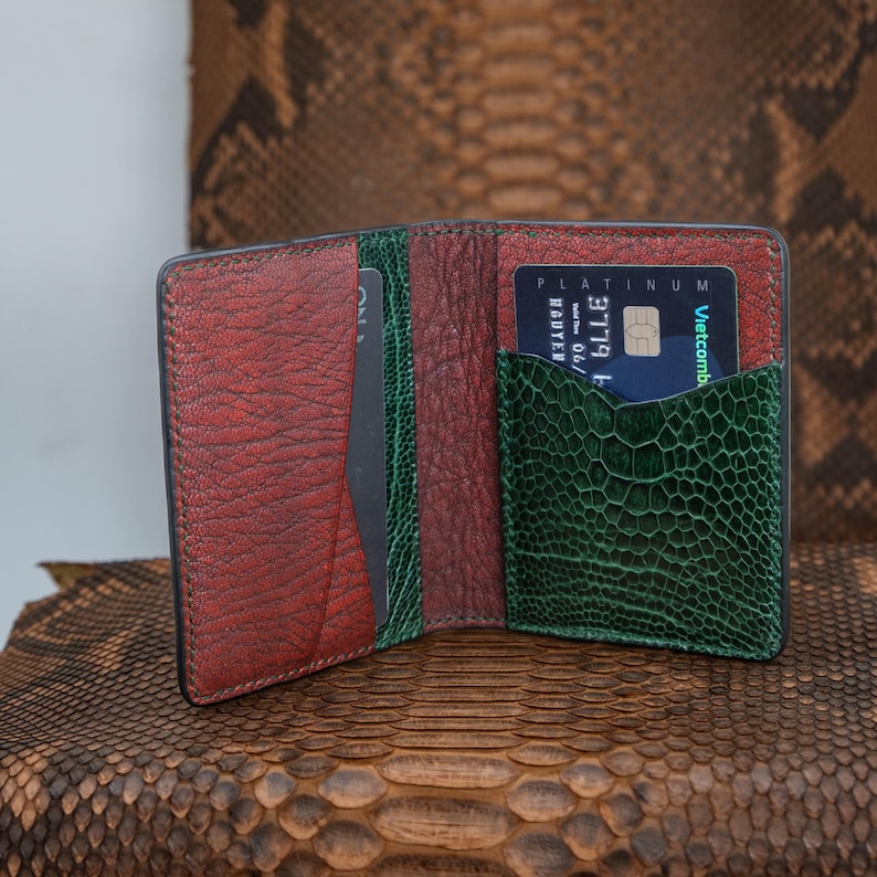 May include: Open leather wallet with a textured, reddish-brown exterior and a green interior pocket. A blue credit card is visible in the pocket. The wallet is open, revealing card slots and a dark interior.