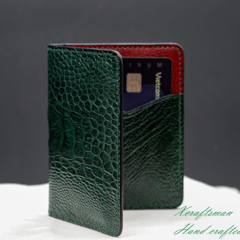 May include: A green leather card holder with a textured, reptile-like pattern. The interior is red, and a blue credit card is visible in a pocket. The wallet is open, displaying its features. The words "Xcraftsman Hand crafted" are in the bottom right corner.