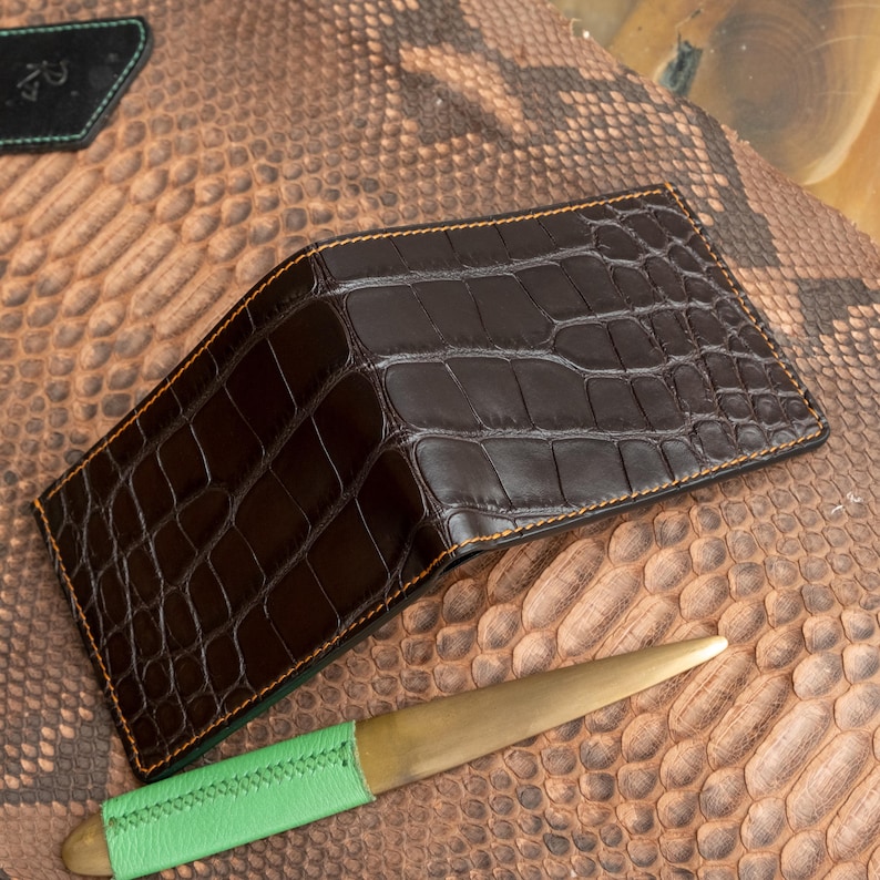 May include: A dark brown leather wallet with a crocodile skin pattern. The wallet has a bi-fold design with orange stitching around the edges. A gold letter opener with a green leather handle is also visible.