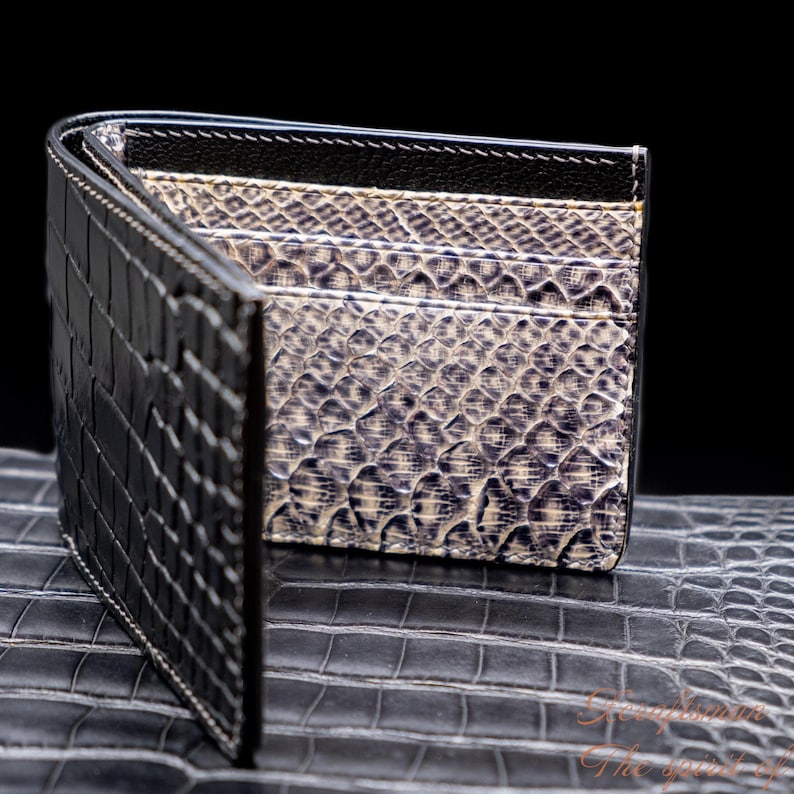 Puede incluir: Una cartera de cuero negra con un patr&oacute;n de cocodrilo en el exterior y un patr&oacute;n de piel de serpiente gris en el interior. La cartera est&aacute; abierta y se encuentra sobre una superficie negra.