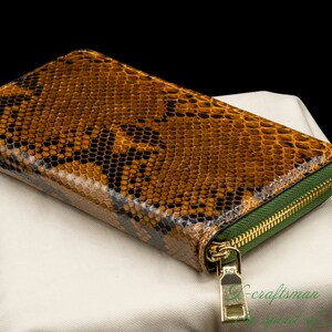 May include: A brown snakeskin wallet with a green zipper and a gold zipper pull. The wallet is lying on a white fabric surface.