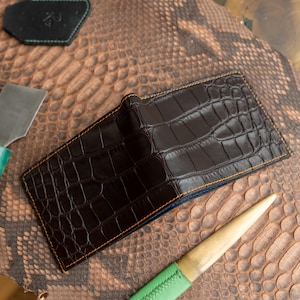 May include: A dark brown leather wallet with a crocodile skin texture. The wallet has orange stitching and a blue interior. The wallet is rectangular and appears to be new. The background is a snakeskin pattern.
