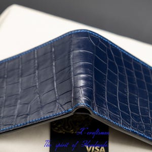 May include: A dark blue alligator skin wallet with a blue stitching detail. The wallet is folded closed and the stitching is visible on the edges.