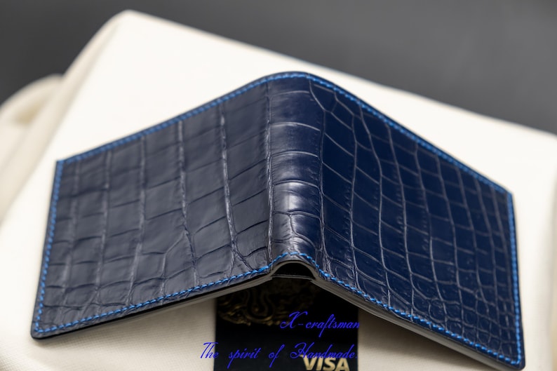 May include: A dark blue alligator skin wallet with a blue stitching detail. The wallet is folded closed and the stitching is visible on the edges.