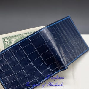 May include: A dark blue alligator skin wallet with a light blue stitch detail. The wallet is open and has cash inside.