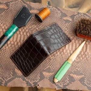 May include: A dark brown leather wallet with a crocodile skin pattern, lying on a snakeskin patterned surface. Other leather working tools include a green and black leather tool, a brush, and a spool of thread.